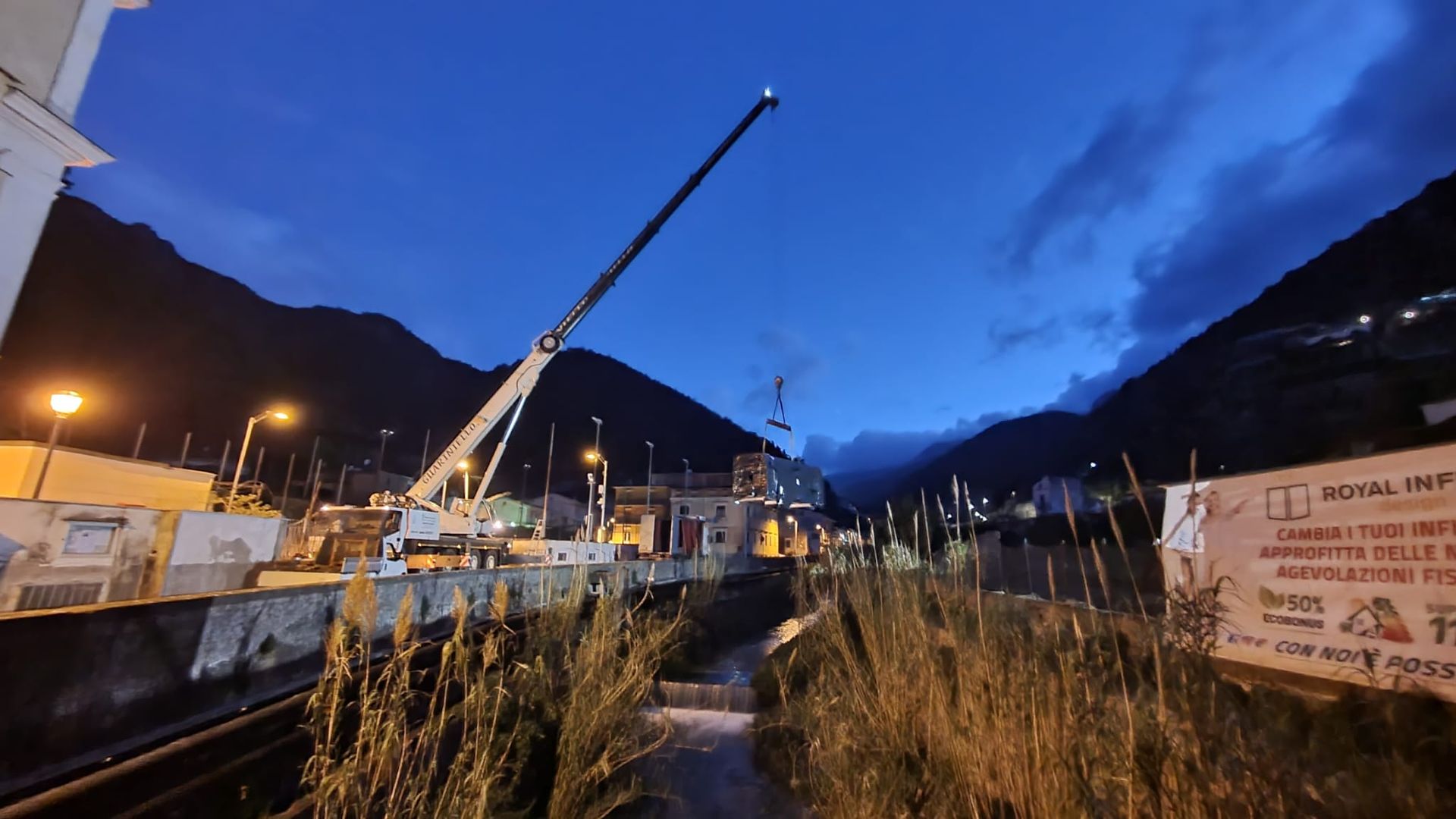 Crepuscolo in un porto di montagna con una gru, barche e luci del molo sotto un cielo azzurro.