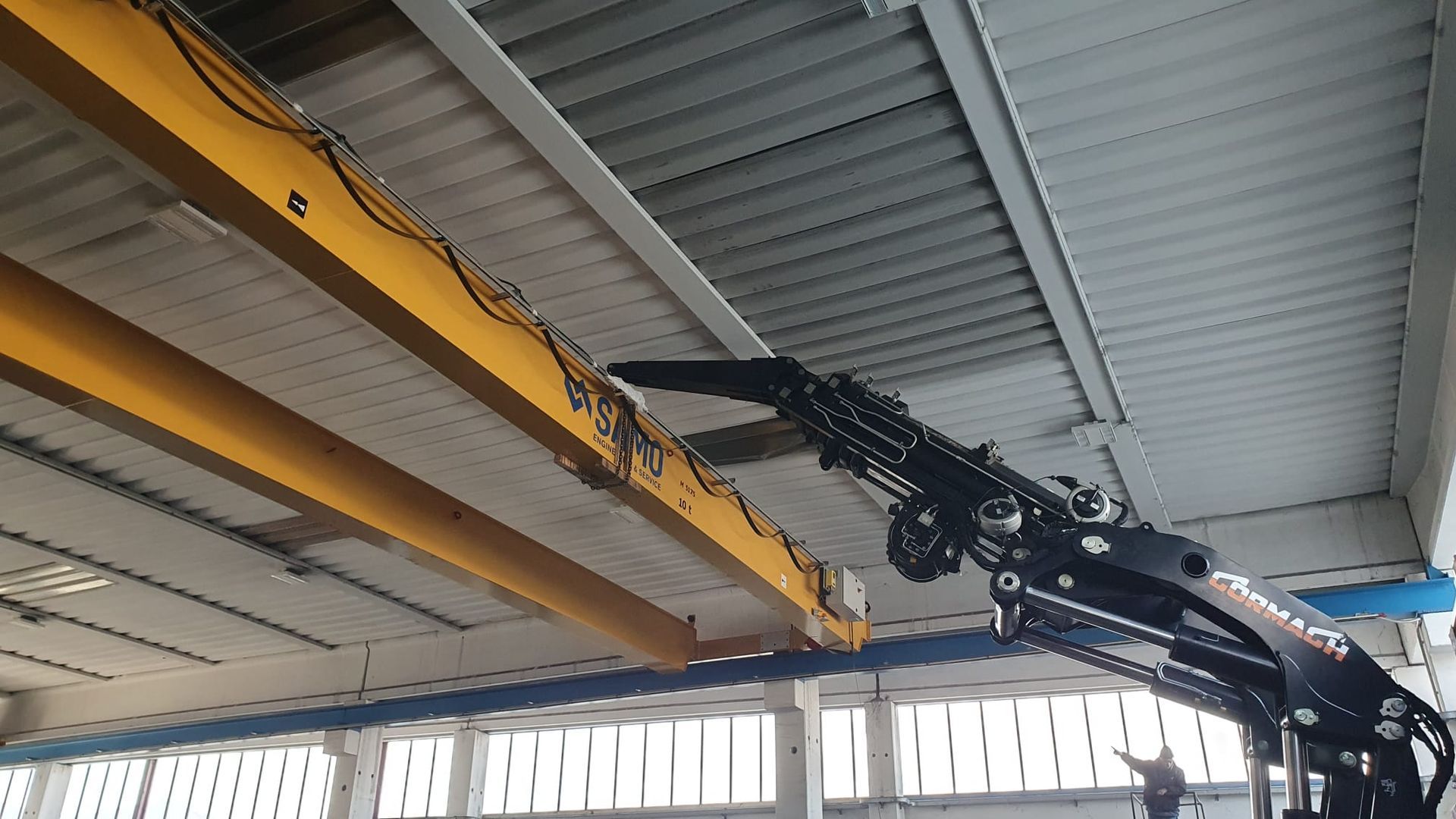 Gru a ponte gialla e braccio idraulico nero all'interno del soffitto di un magazzino industriale.