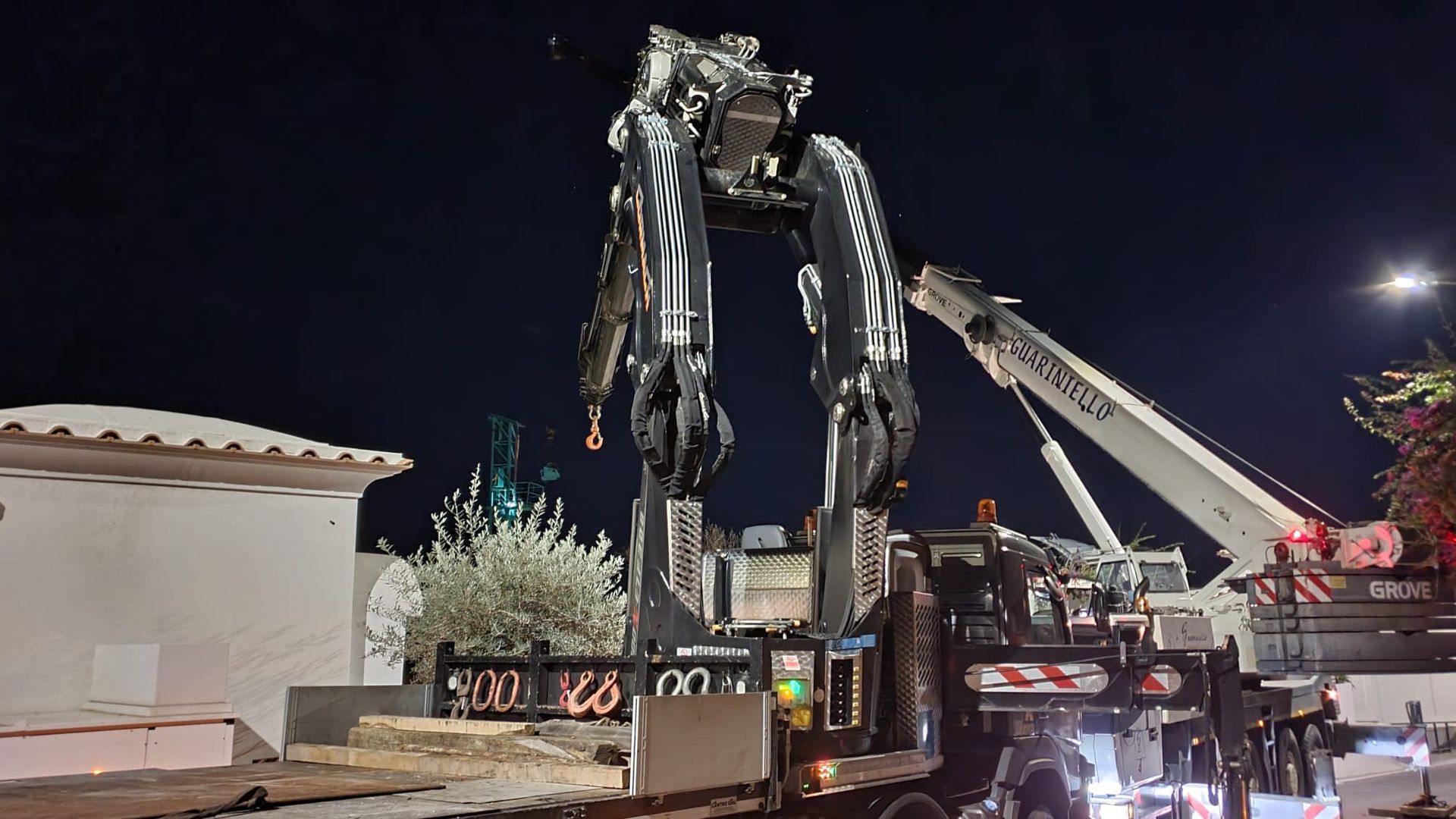 Di notte, una gru solleva la fusoliera danneggiata di un aereo vicino a un edificio illuminato e a un camion dei pompieri.