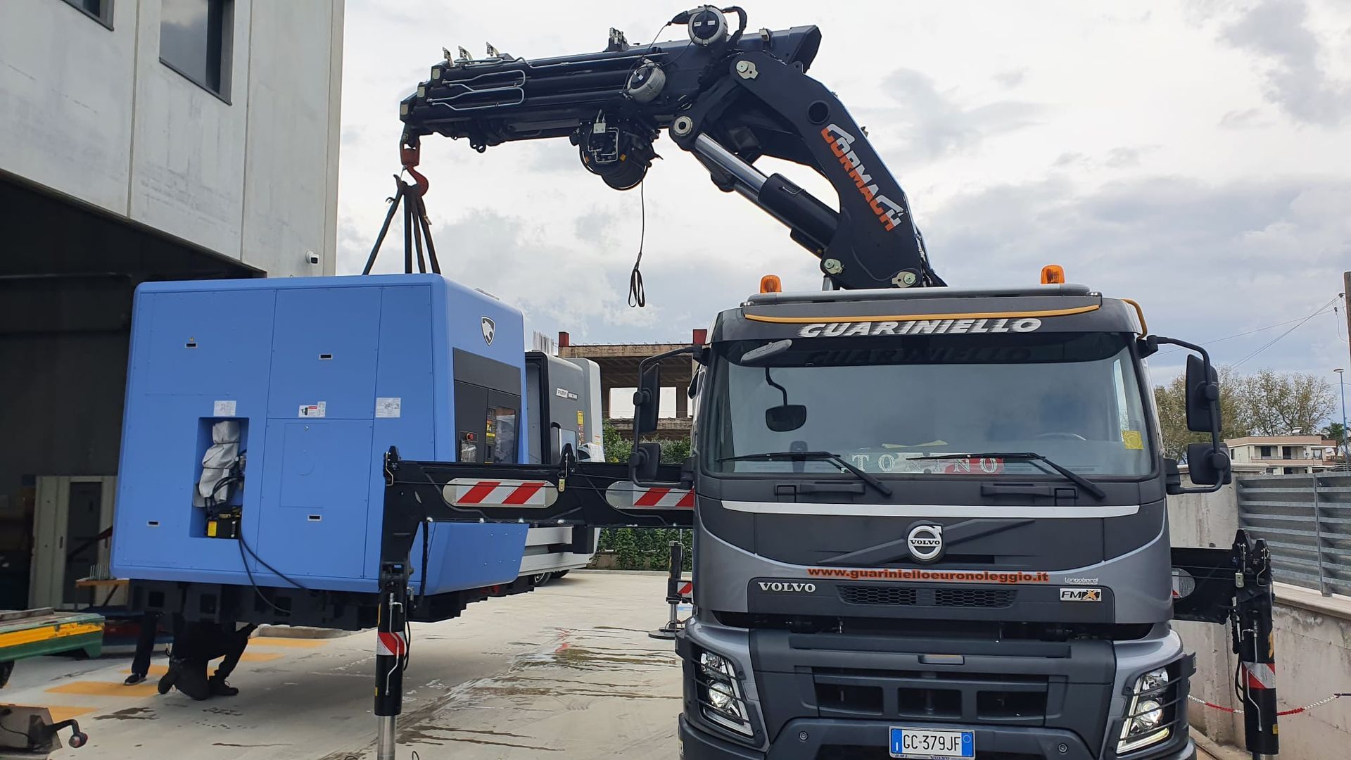 Un camion con gru solleva un grande macchinario industriale blu accanto a un edificio in un cantiere.