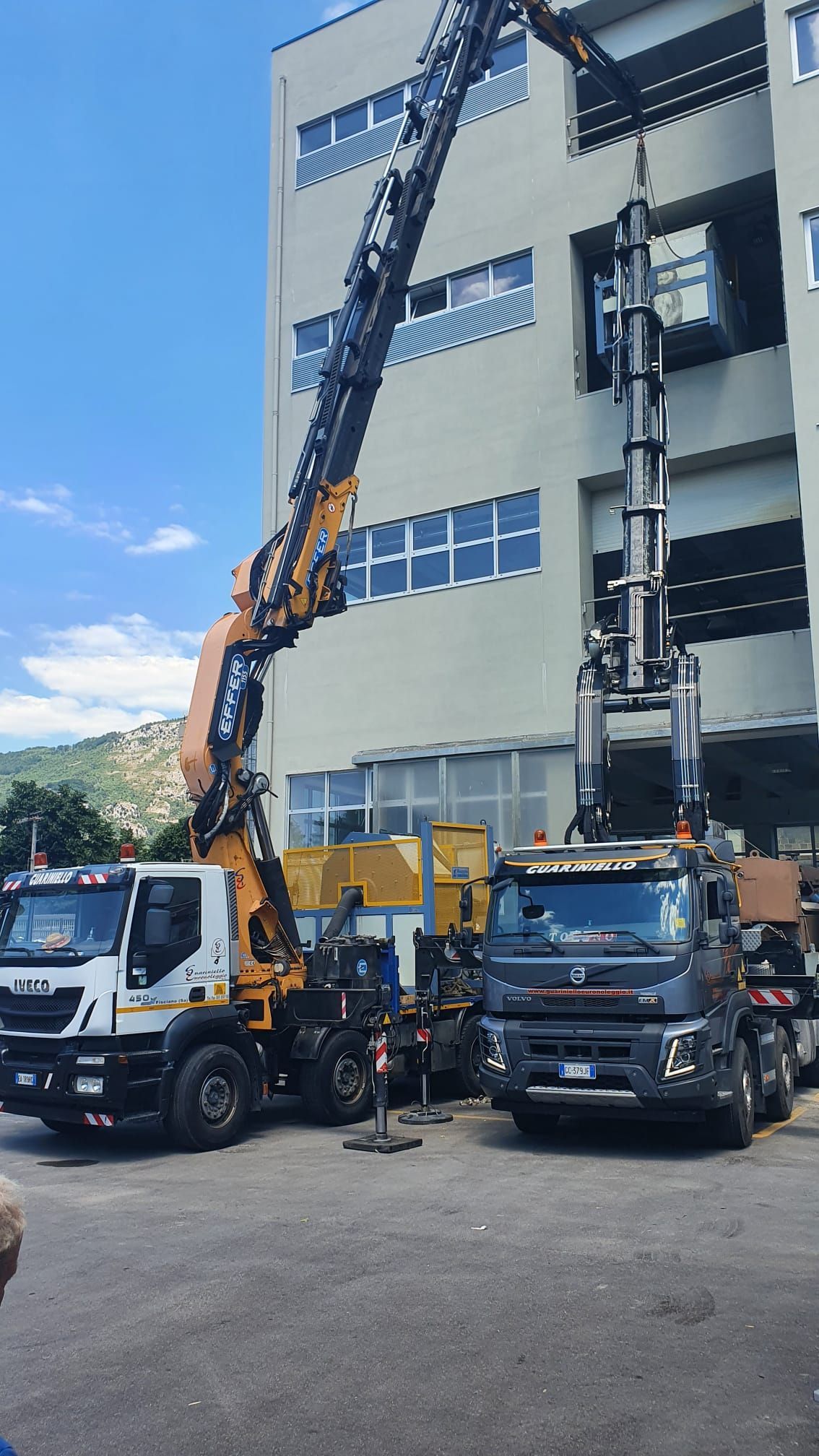 Una gru da costruzione solleva materiali accanto a un edificio a più piani, con camion parcheggiati al di sotto.
