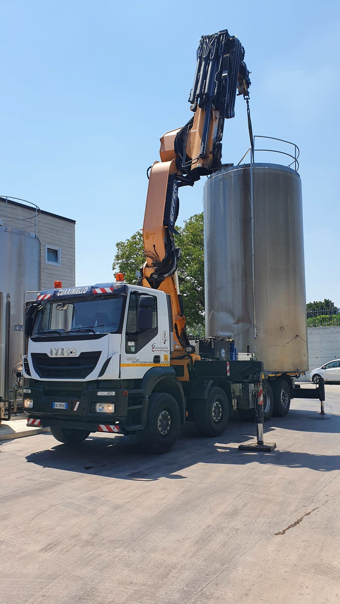 Un camion bianco con gru solleva un grande serbatoio cilindrico argentato in un'area industriale.