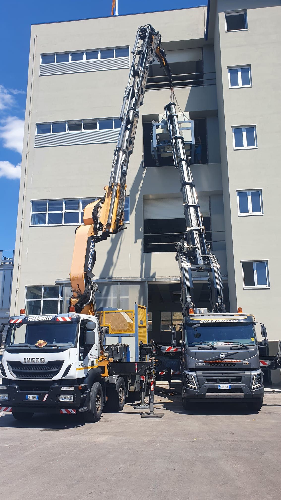 Due gru sollevano attrezzature contro un edificio grigio, con dei camion parcheggiati davanti.