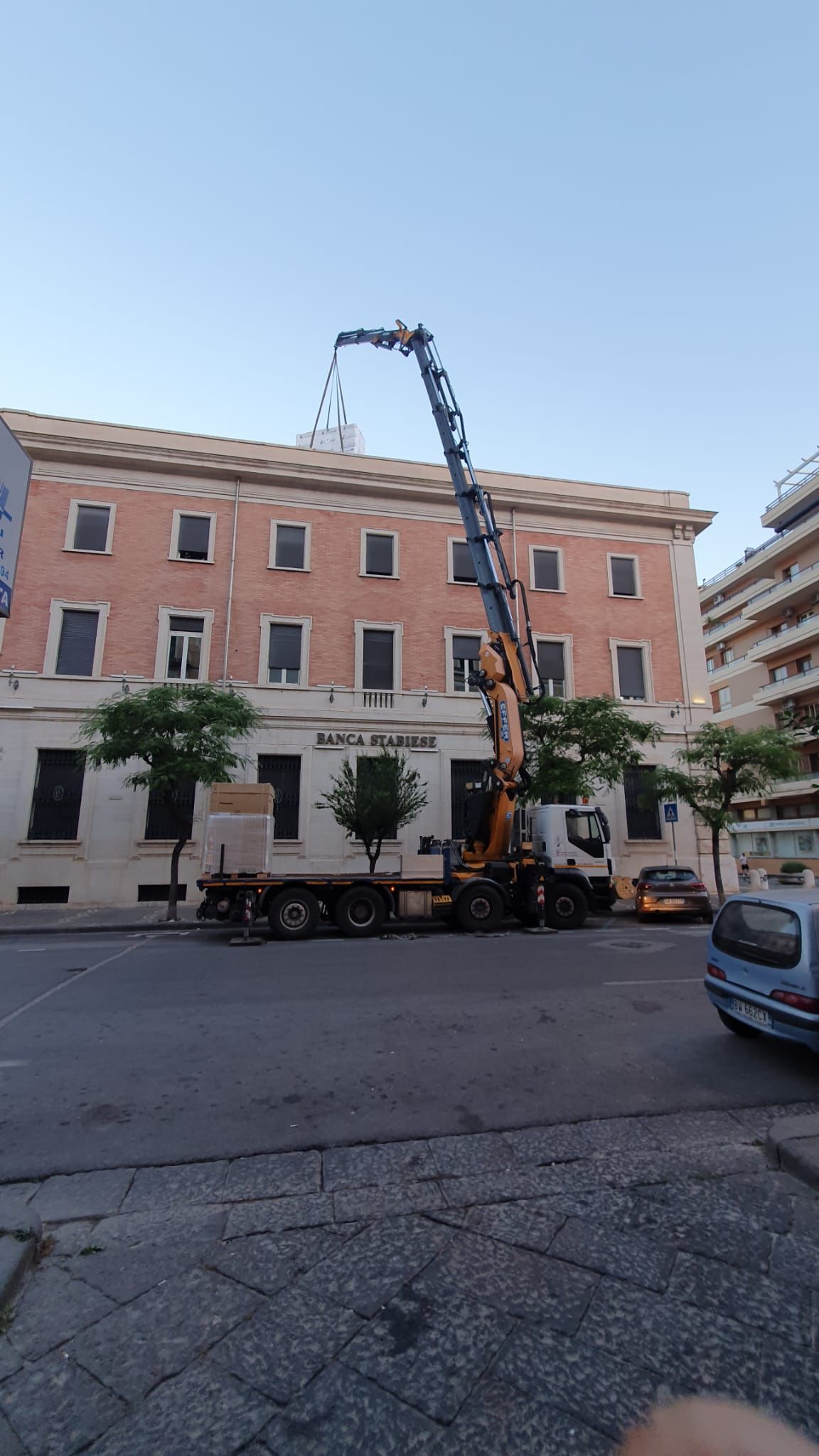 Un camion per la potatura degli alberi con il braccio sollevato si trova su una strada cittadina accanto a un edificio in mattoni al crepuscolo.