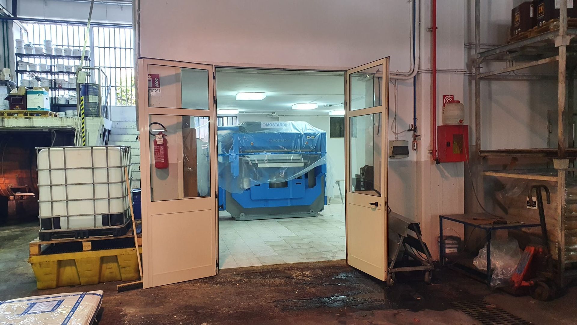 Ingresso di un magazzino industriale con doppie porte aperte e un macchinario blu all'interno, circondato da pallet e attrezzature.