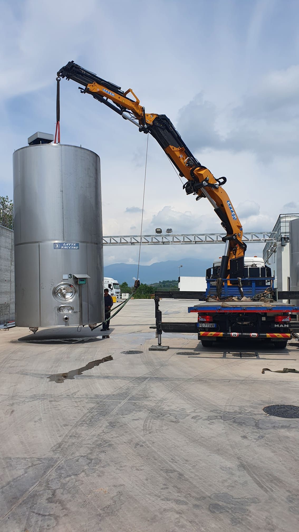 Una gru arancione solleva una lunga trave sopra un grande serbatoio argentato in un'area industriale.