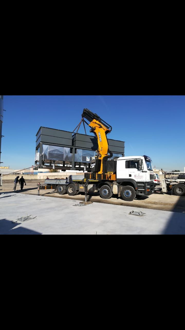 Un camion bianco con una gru gialla solleva travi d'acciaio in un cantiere edile sotto un cielo azzurro e limpido.