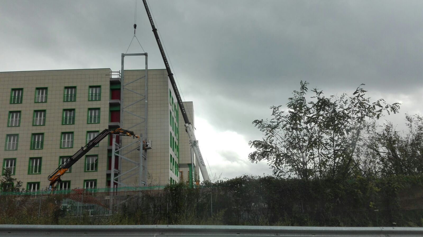 Via cittadina piovosa con un edificio color beige, alberi e una gru edile sotto un cielo grigio.