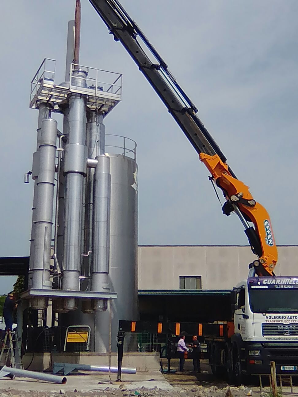Una gru solleva un alto serbatoio industriale argentato accanto a un camion bianco in un cantiere.