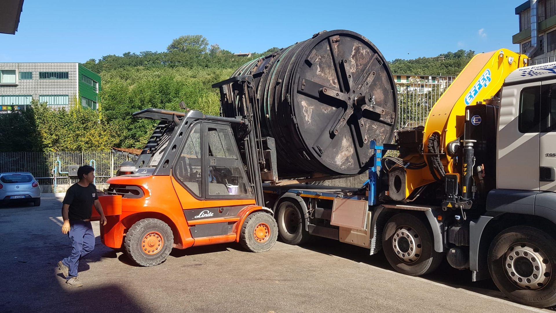 Carrello elevatore arancione che carica una grande bobina di cavo nera su un camion a pianale all'aperto.