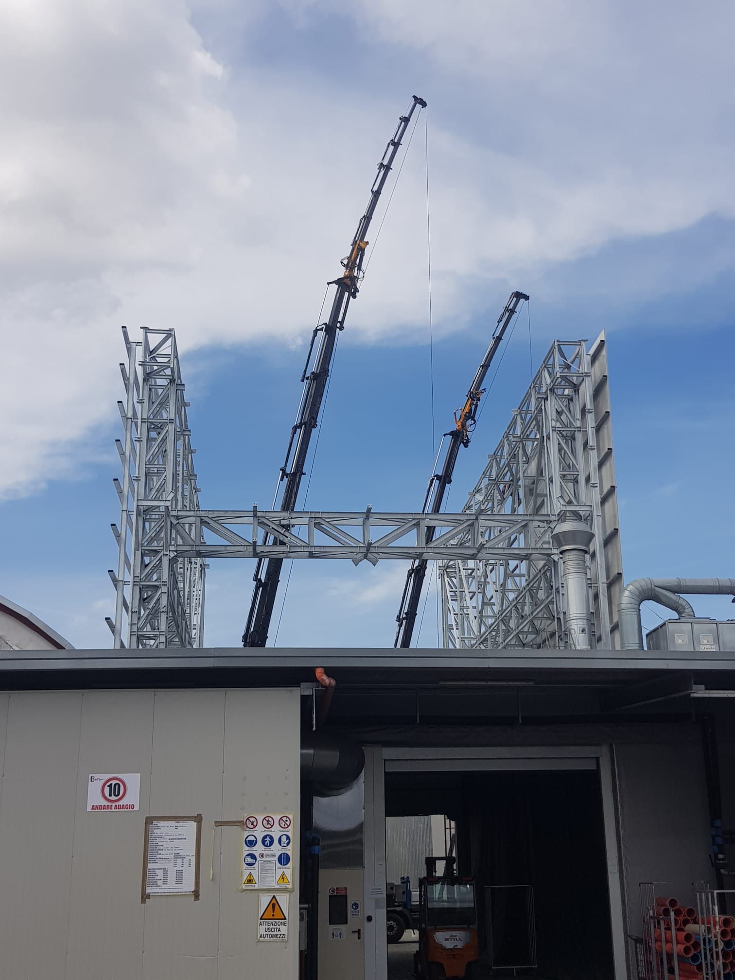 Cantiere industriale con struttura in acciaio e due gru sullo sfondo di un cielo nuvoloso.