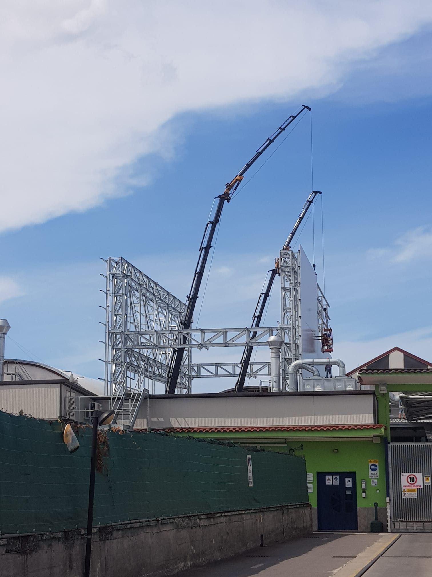 Gru da costruzione sopra un cantiere con struttura in acciaio e recinzione verde sotto un cielo nuvoloso.