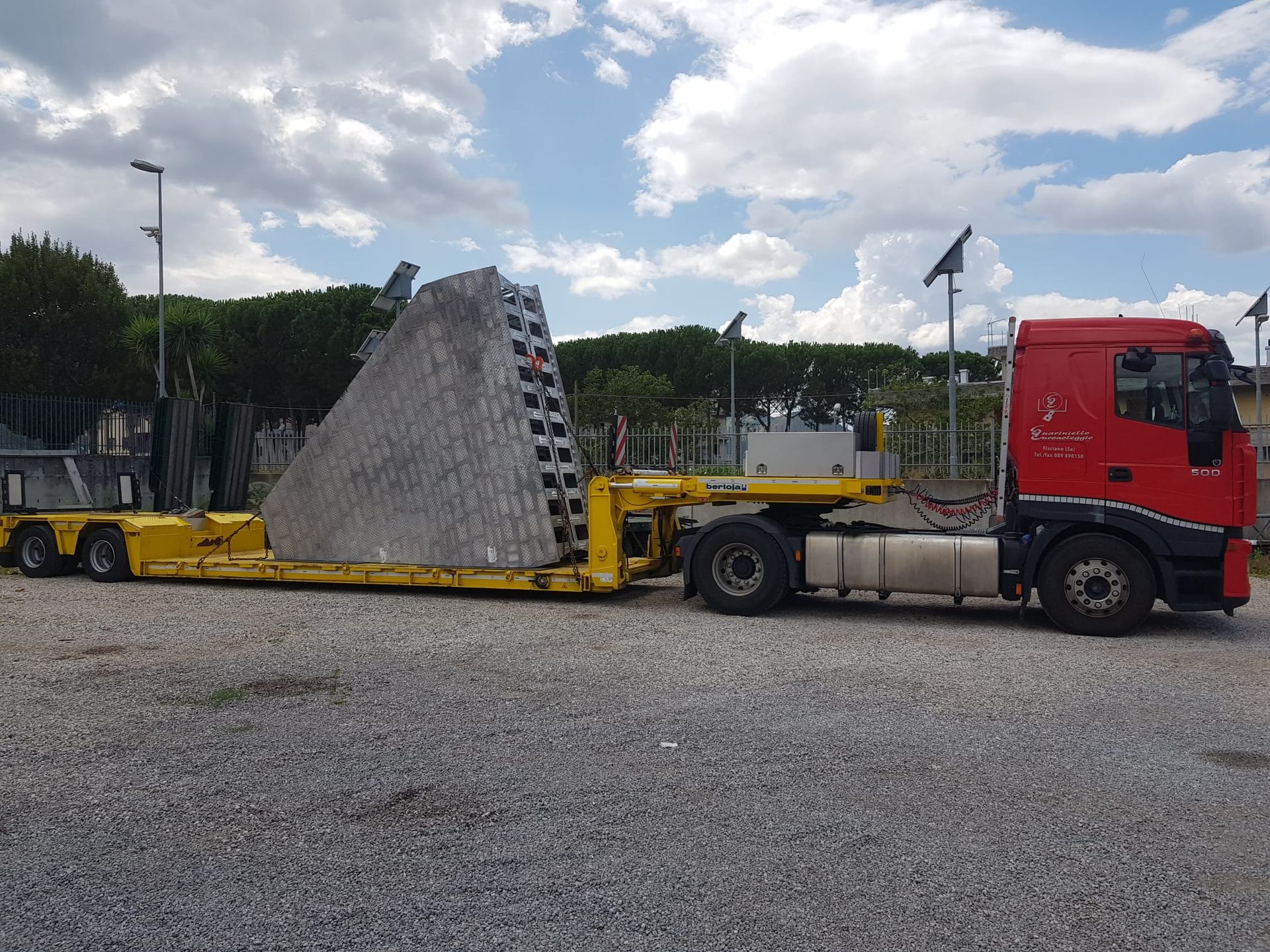 Un camion rosso con semirimorchio trasporta un grosso blocco di cemento su un rimorchio a pianale giallo in un piazzale ghiaioso.