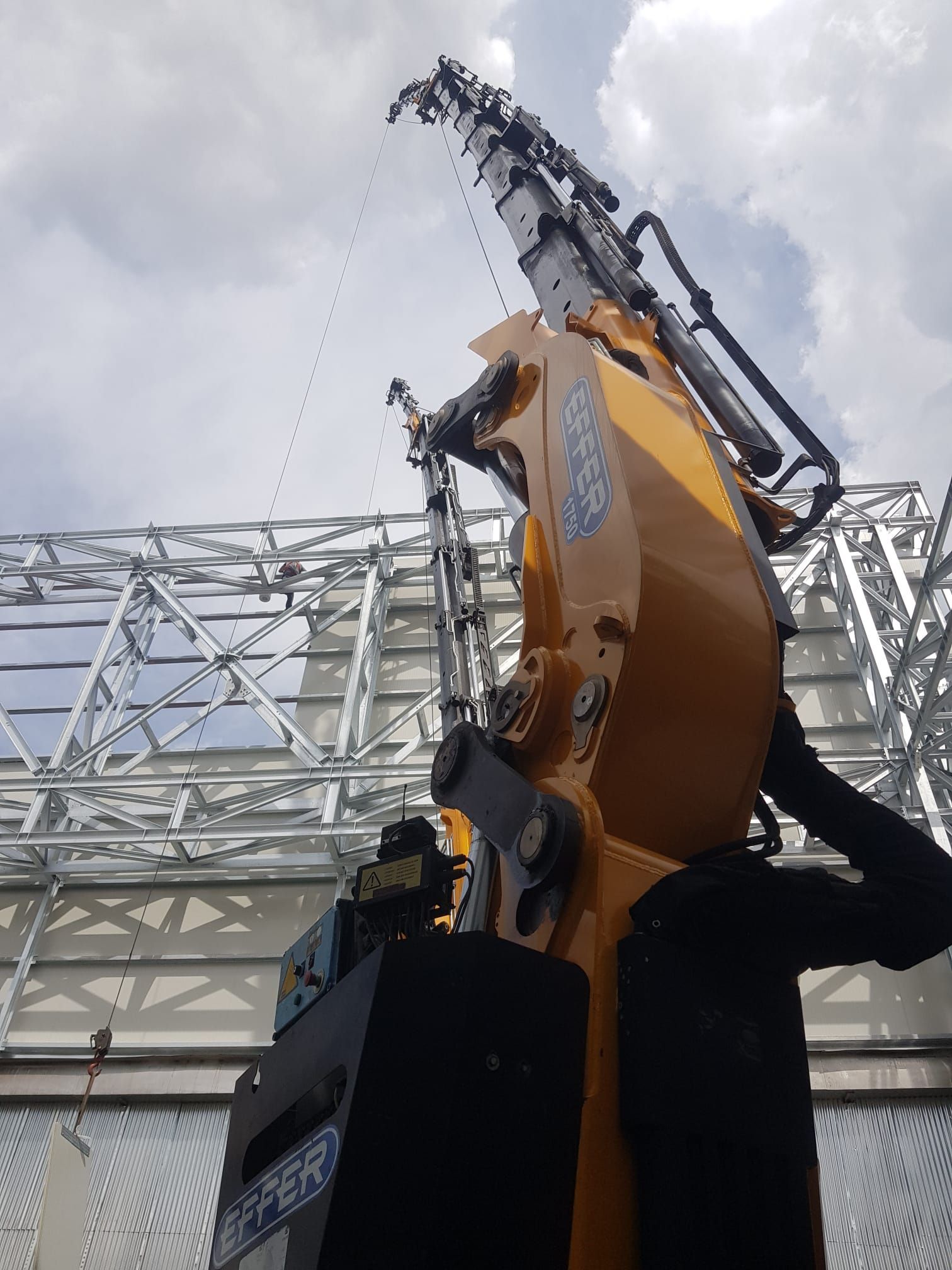 Grande macchina da costruzione gialla con braccio sollevato accanto a una struttura metallica sotto un cielo nuvoloso.