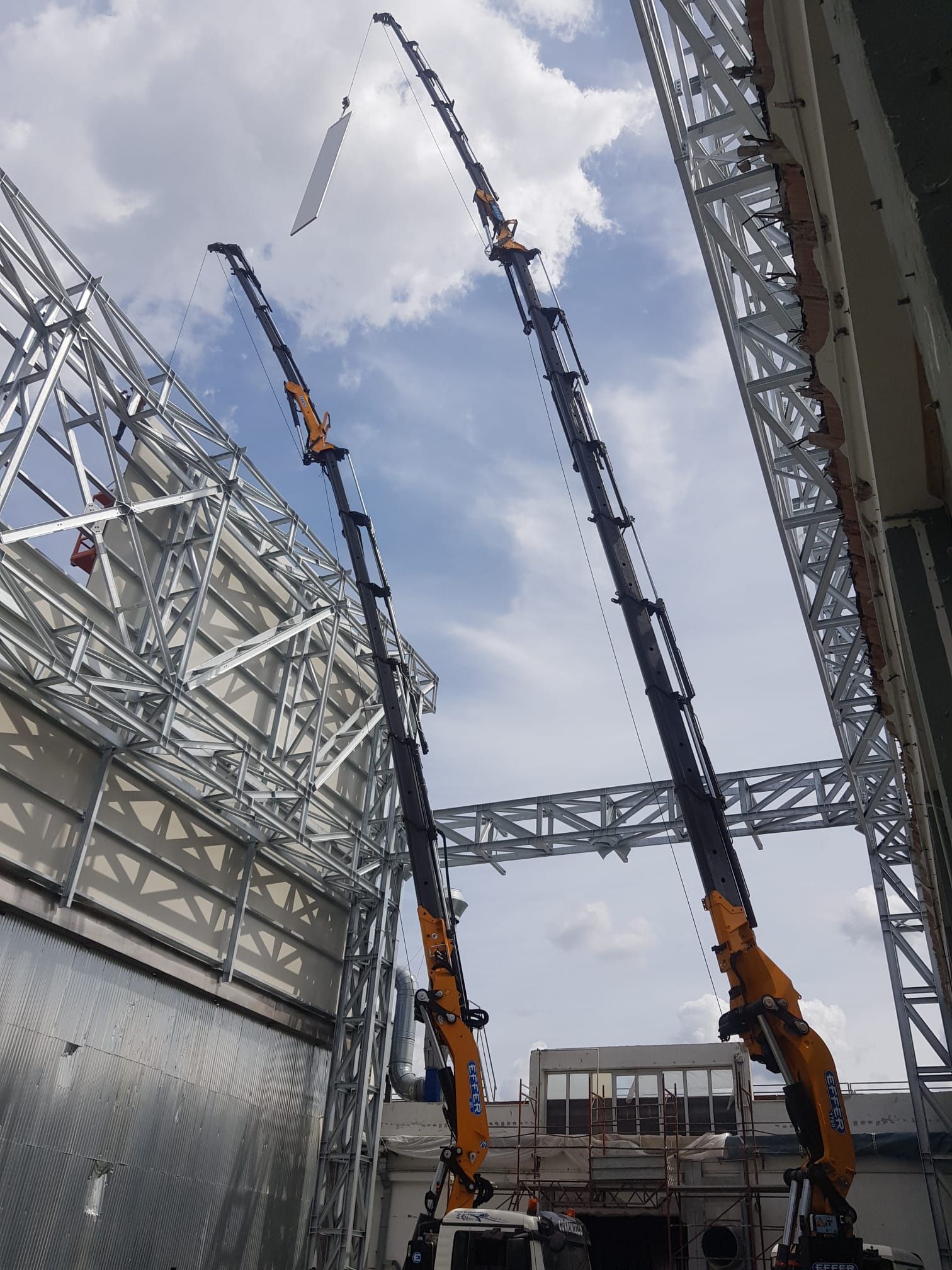 Cantiere edile con due alte gru accanto a un edificio in metallo sotto un cielo nuvoloso.