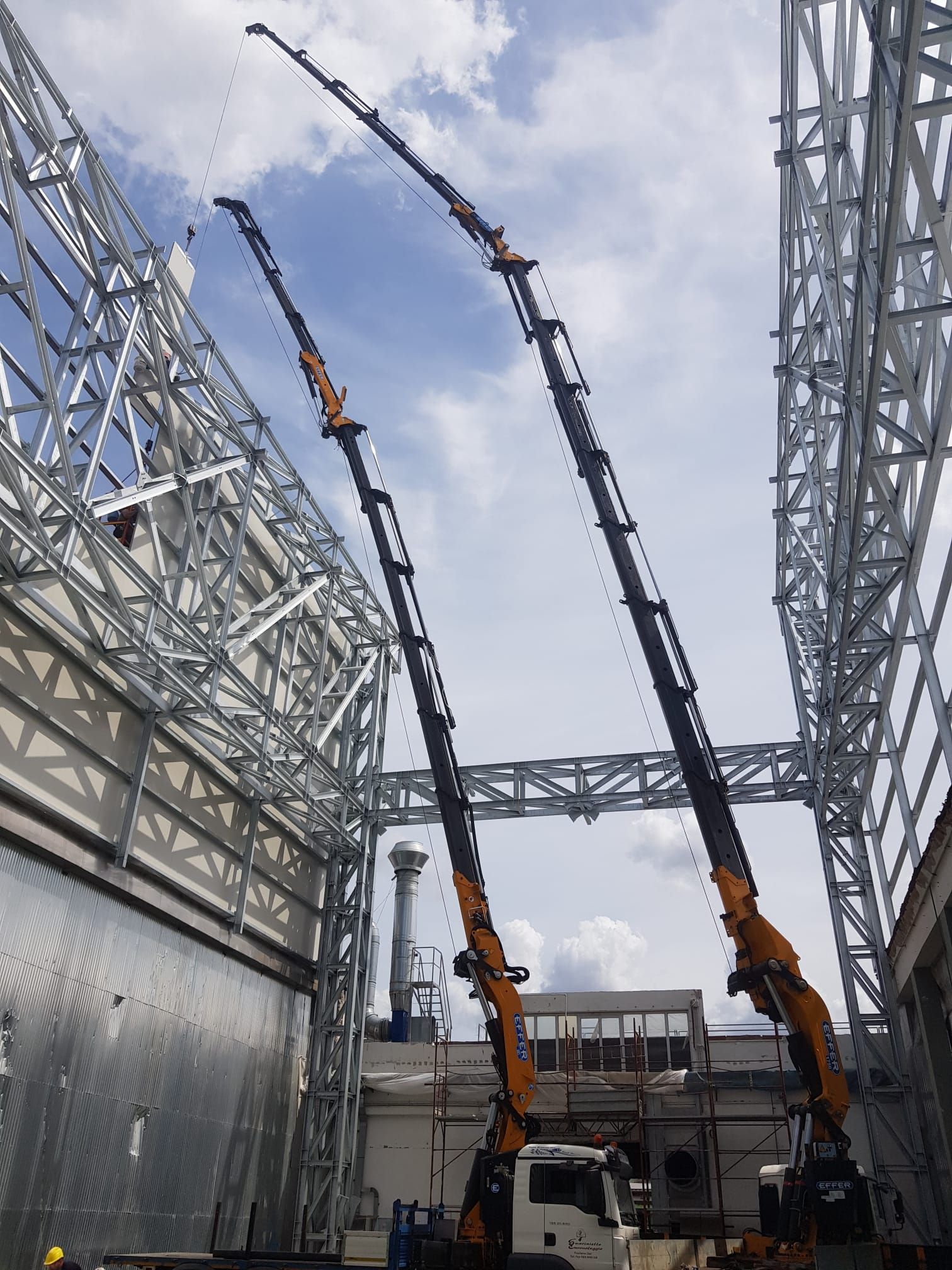 Due grandi gru sollevano travi d'acciaio tra strutture industriali in un cantiere.