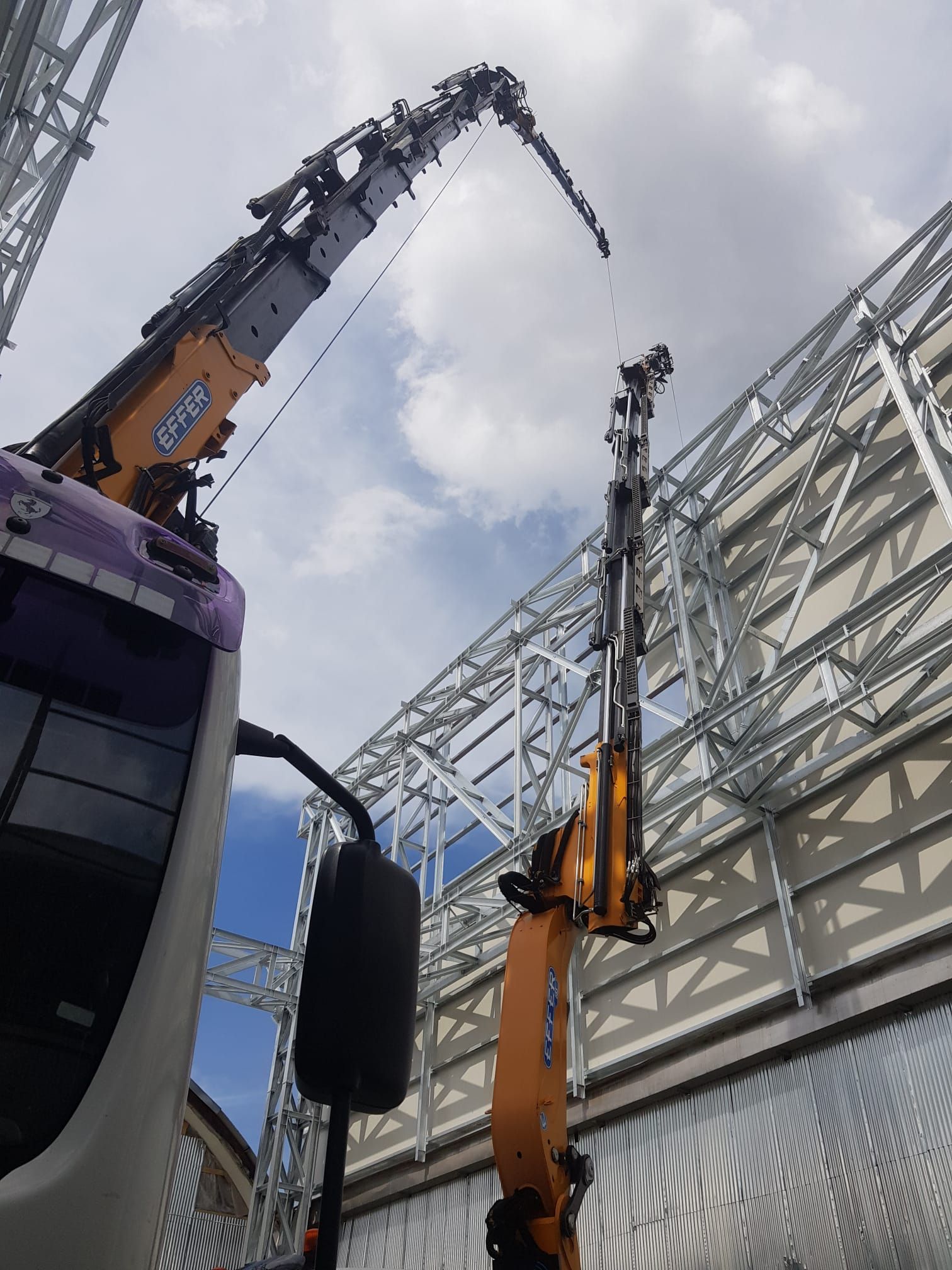 Due gru da costruzione arancioni sollevano una struttura metallica contro un cielo nuvoloso.