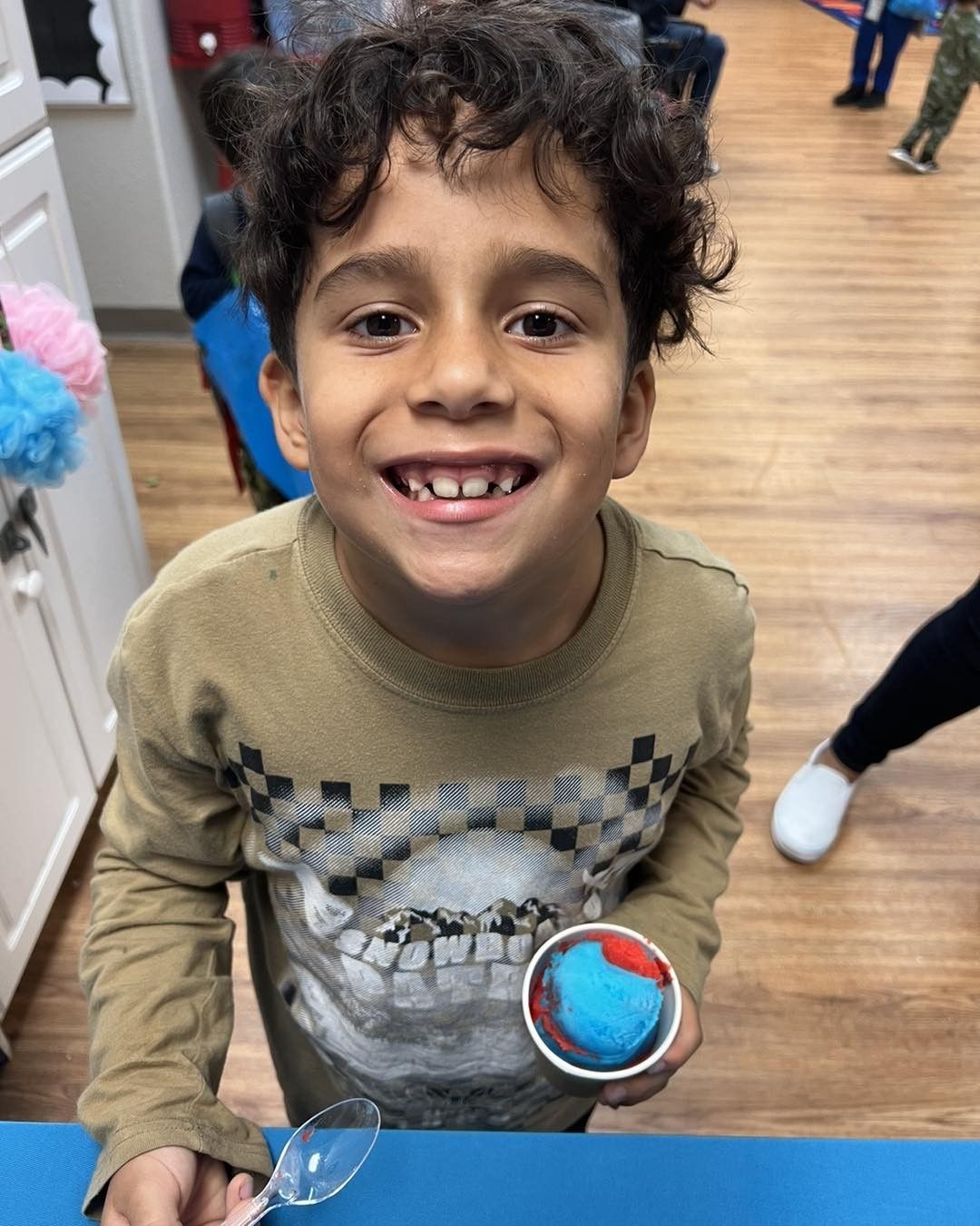 Boy smiles, holding a cup of blue and red frozen treat, inside a room.