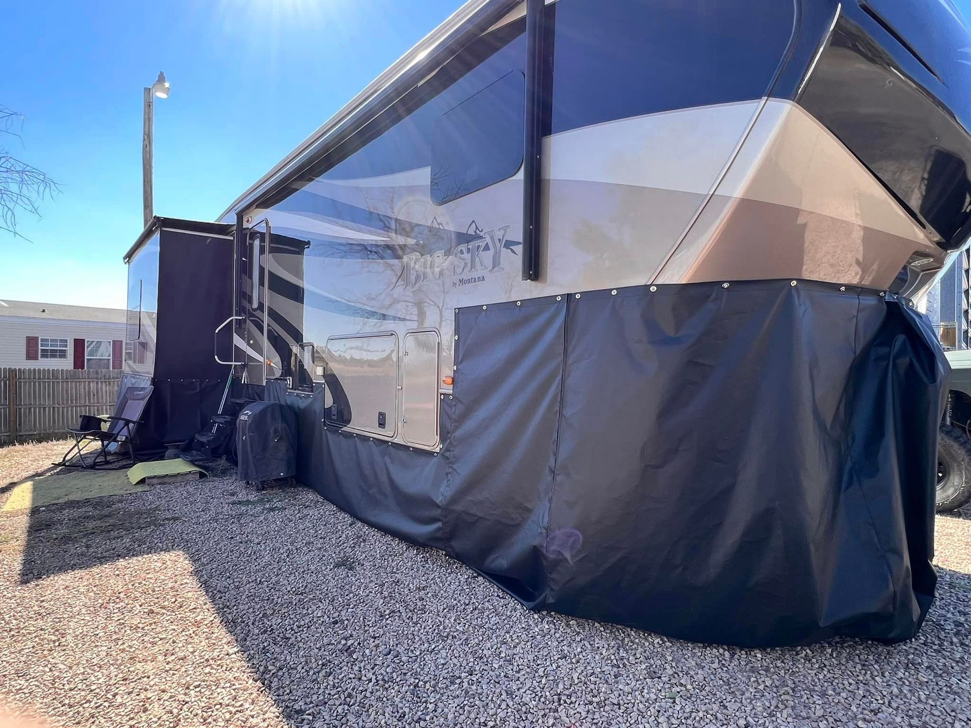 Blue-and-white RV with extended awning and side panels parked on gravel beside a small building.