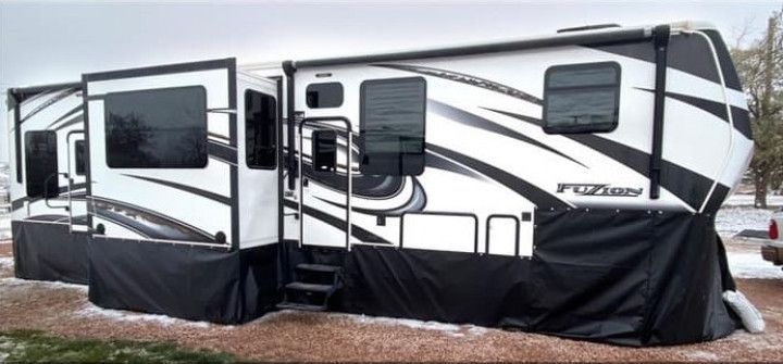 White and black RV trailer with extended slide-outs and black skirting parked on muddy ground.