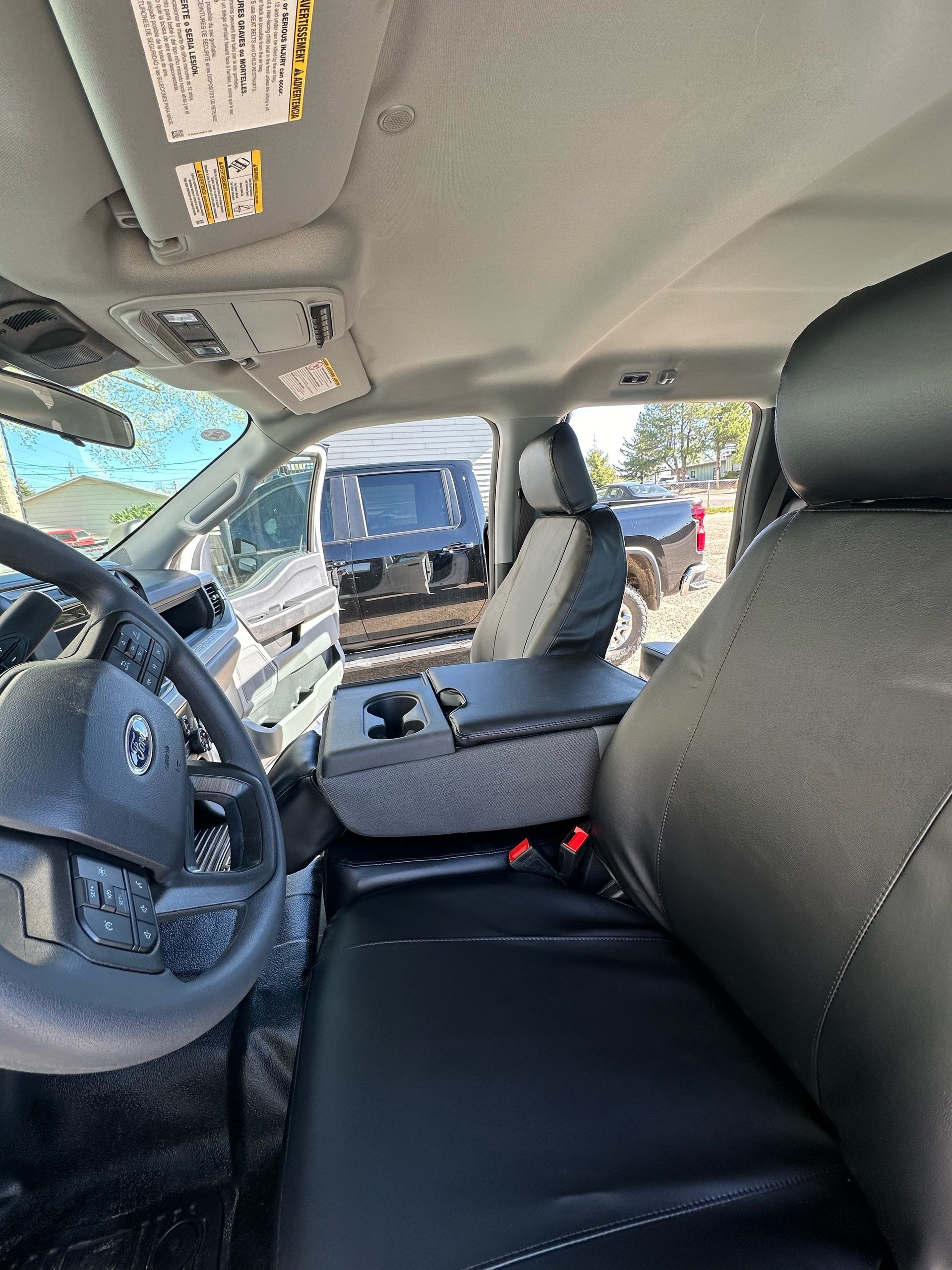 Inside a vehicle cabin with a steering wheel, dashboard, black seats, and the door open to a parking lot.