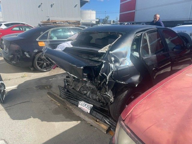 A black car with severe rear-end damage in a parking lot; other vehicles visible.