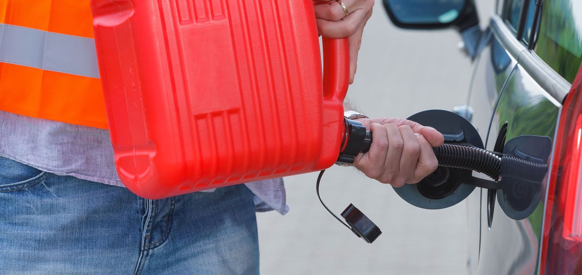 A person wearing a high-visibility orange vest pours fuel from a red plastic gas can into a car's fuel tank.