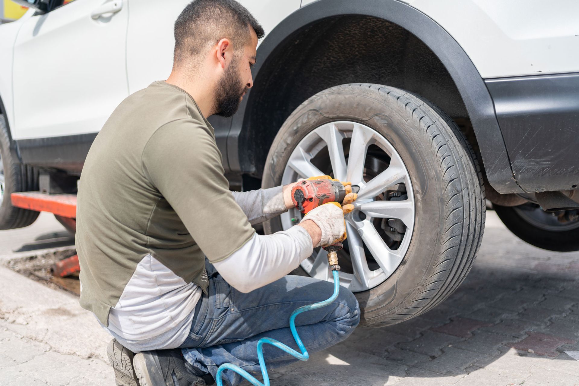 Man kneeling, using an impact wrench to remove lug nuts from a car tire, outdoors. Man kneeling, using an impact wrench to remove lug nuts from a car tire, outdoors.