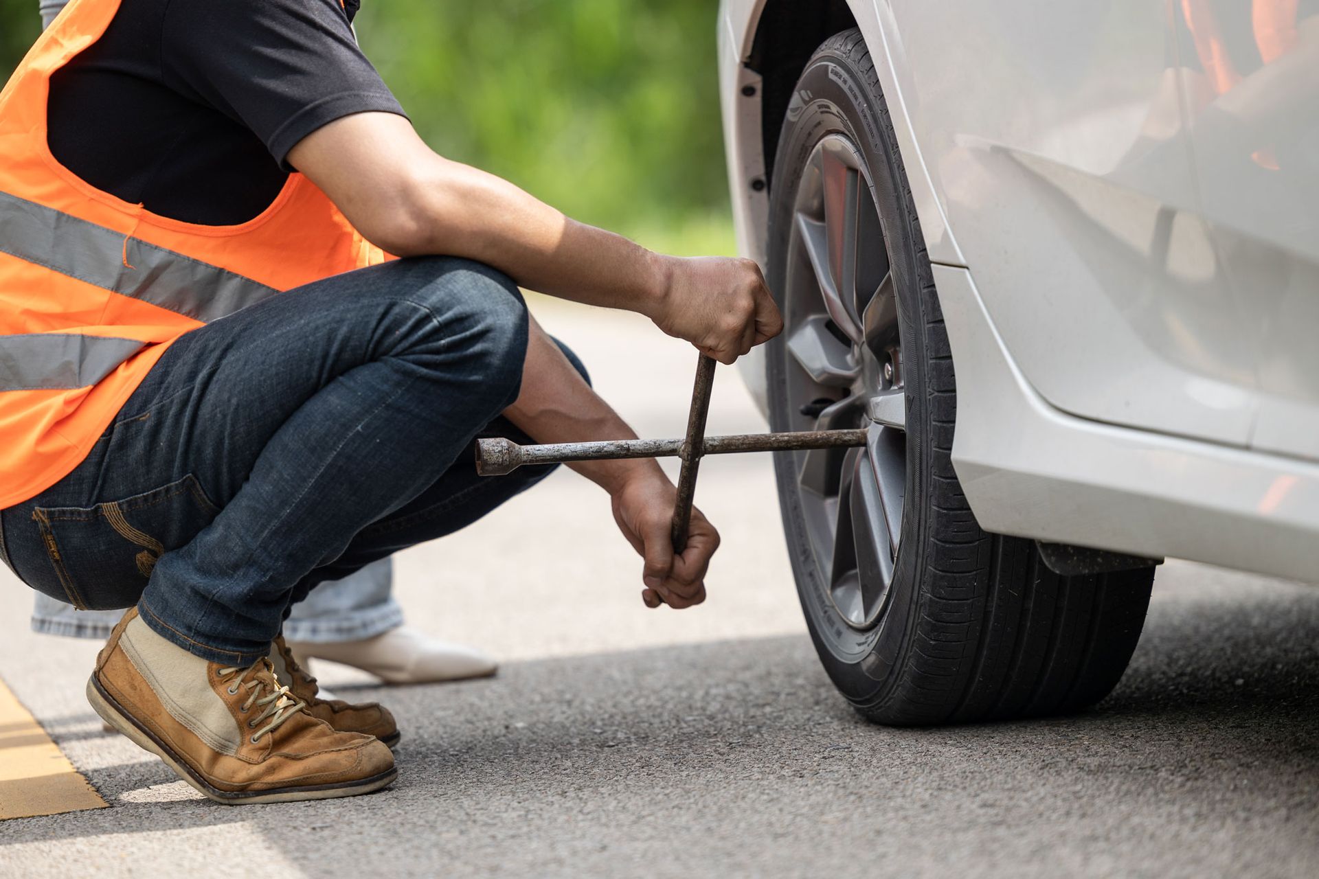 Person in an orange vest changing a flat tire on a white car with a lug wrench on the side of the road. Person in an orange vest changing a flat tire on a white car with a lug wrench on the side of the road.