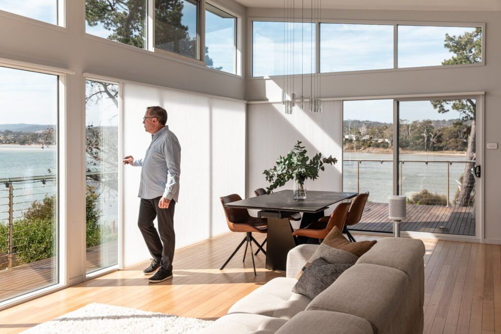 A Man is Standing in a Living Room Looking Out of a Window — TT Shutters & Shading In Ballina, NSW