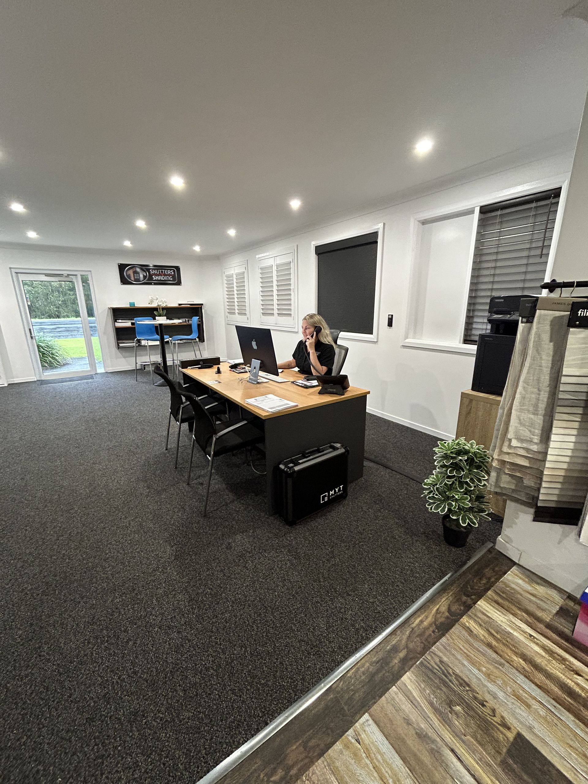 A Woman is Sitting at a Desk in an Office Talking on a Cell Phone — TT Shutters & Shading In Ballina, NSW