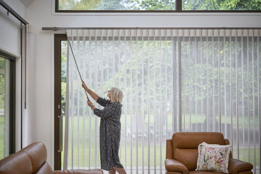 A Woman is Pulling Up Vertical Blinds in a Living Room — TT Shutters & Shading In Ballina, NSW
