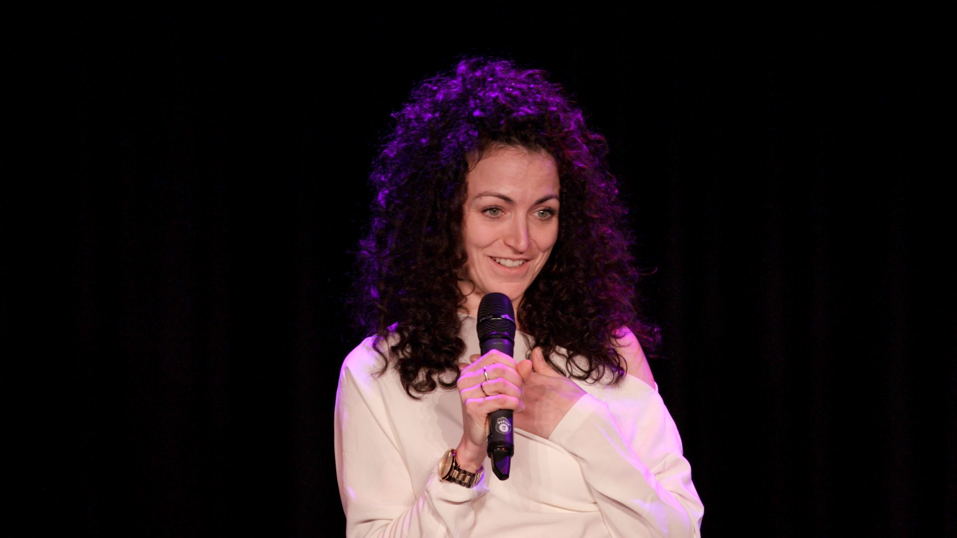 A person with dark curly hair wearing a white top stands on a dark stage, holding a microphone and smiling.