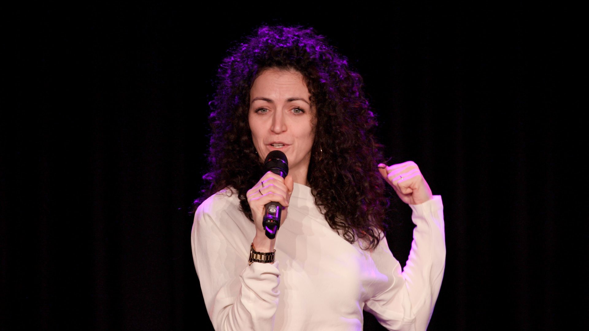 A person with dark curly hair holding a microphone and gesturing on stage against a dark background with pink lighting.