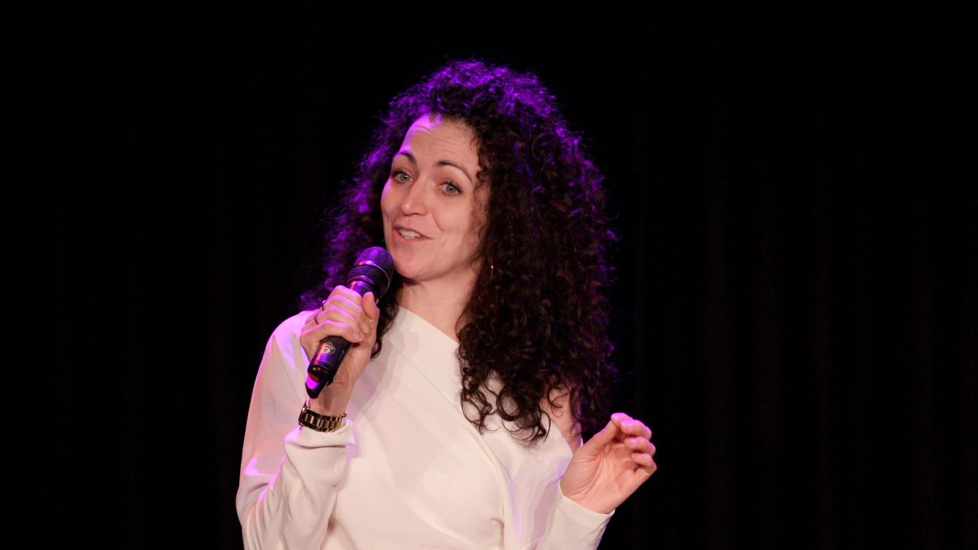 A person with curly dark hair wearing a white top stands against a dark background, speaking into a handheld microphone.