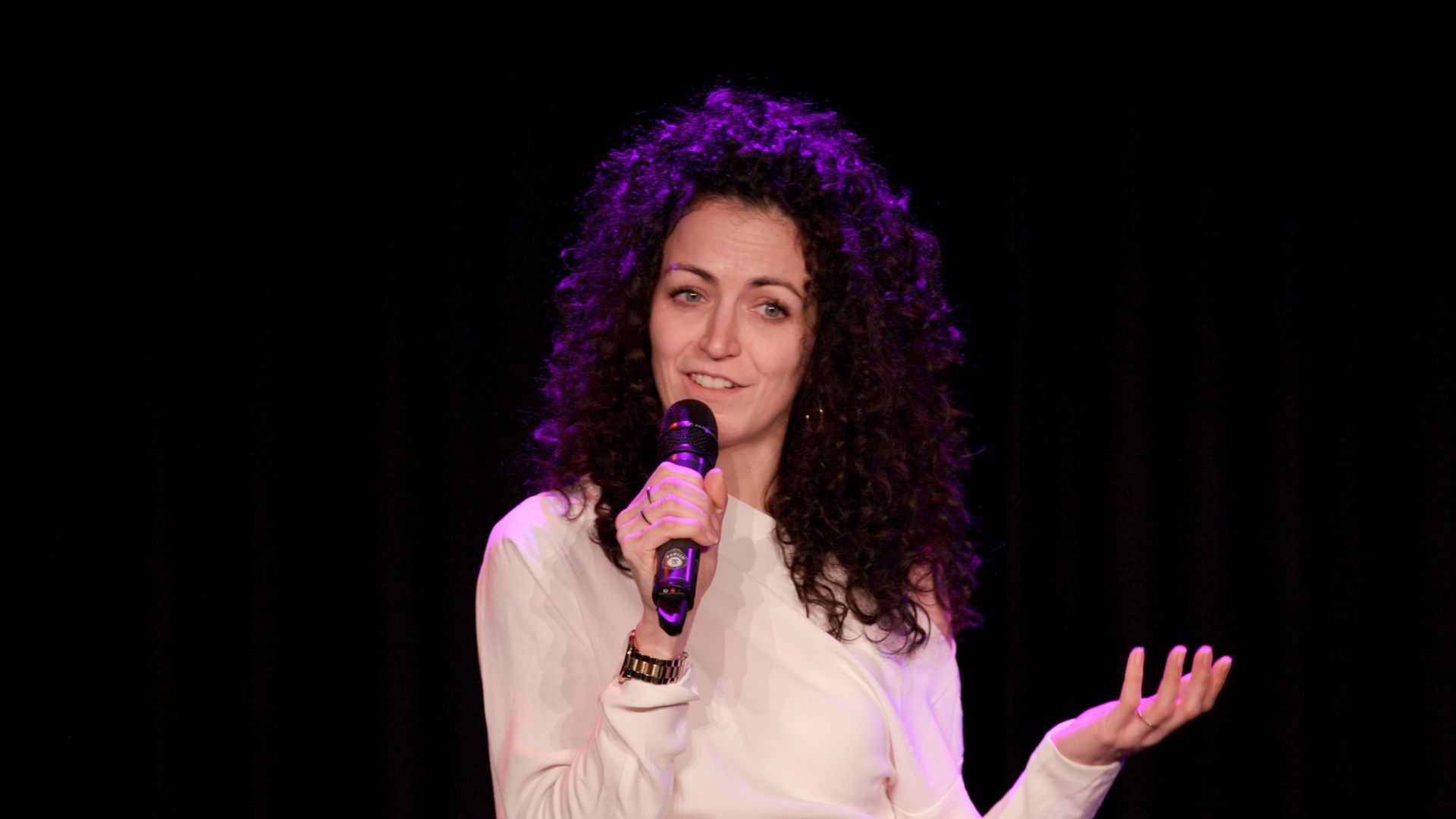A person with curly dark hair wearing a white top speaks into a microphone on a stage with a dark background.