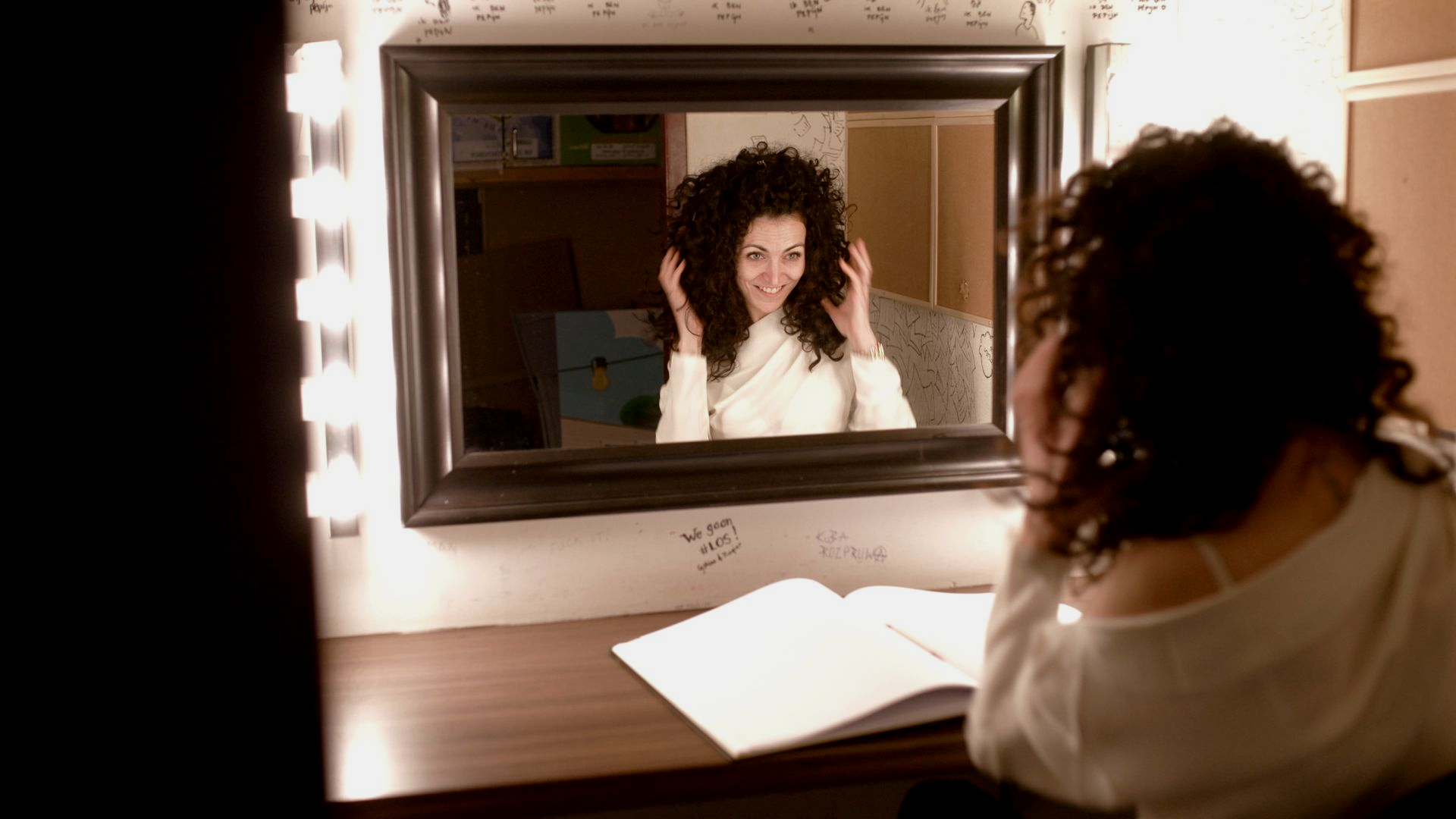 A person with curly hair sits at a vanity with glowing lights, looking at their reflection in a framed mirror.