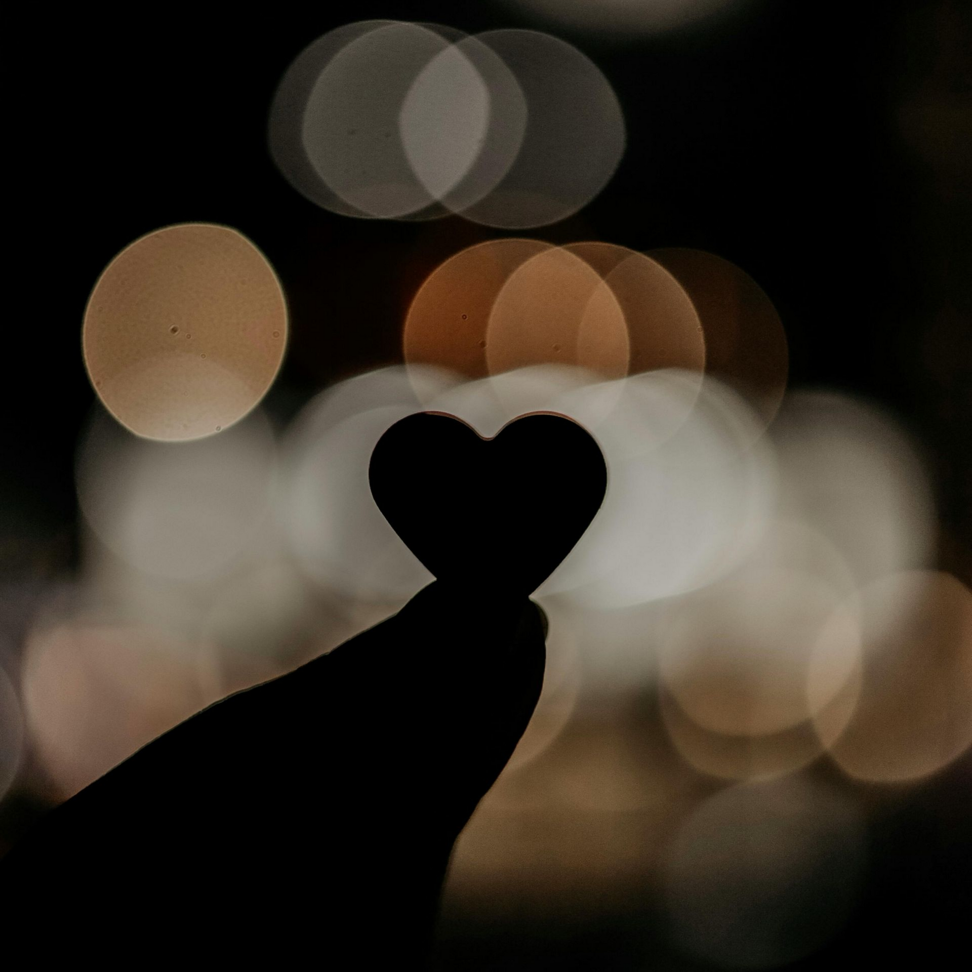 A silhouette of a hand holding a small heart shape against a blurred background of warm, glowing bokeh lights.