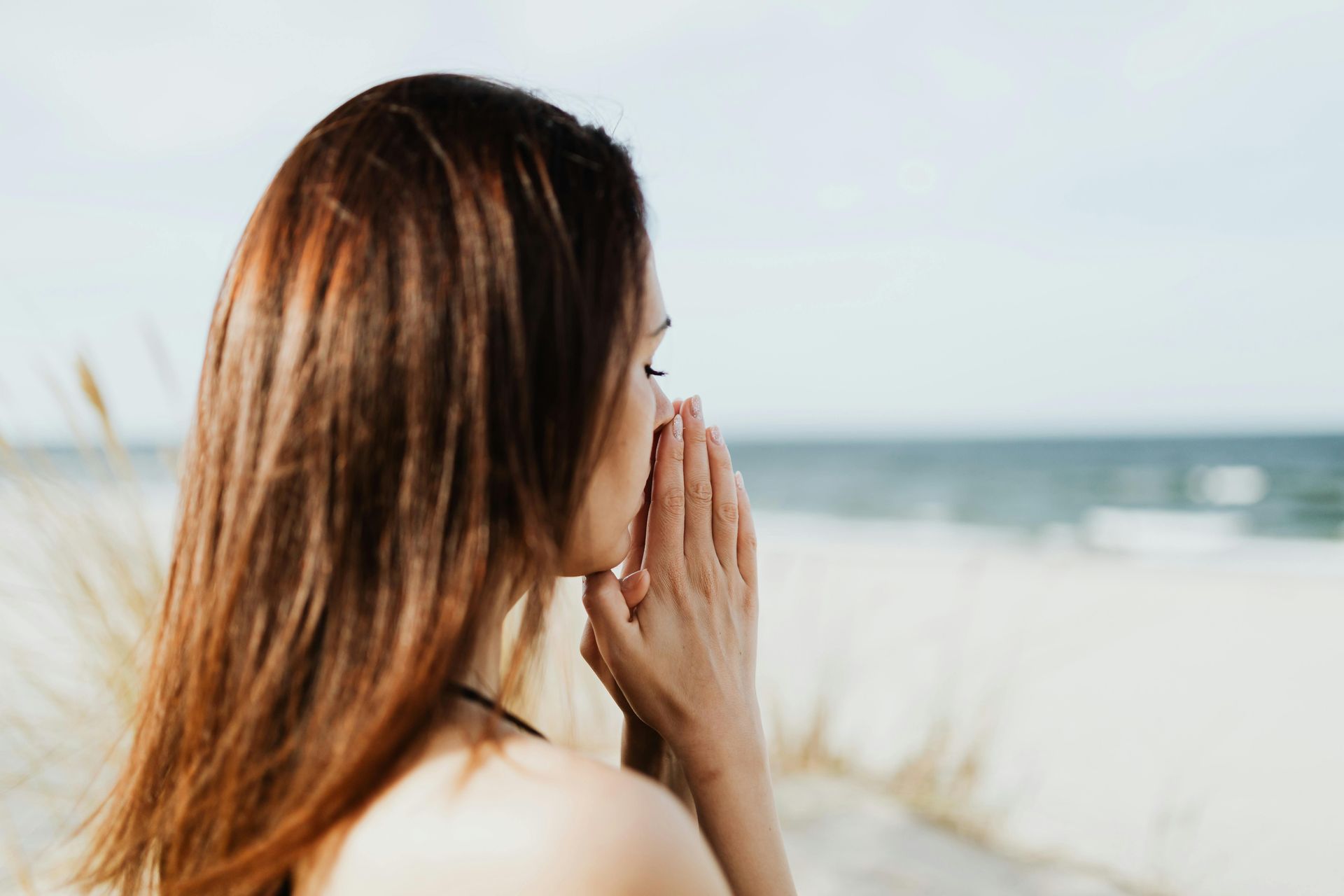 A person with long brown hair stands on a beach, looking toward the ocean with hands held together near their face.