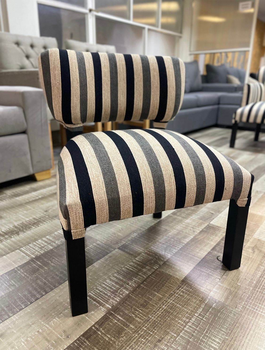 Una silla pequeña sin brazos, con asiento y respaldo tapizados a rayas negras, grises y beige, y patas de madera negra.