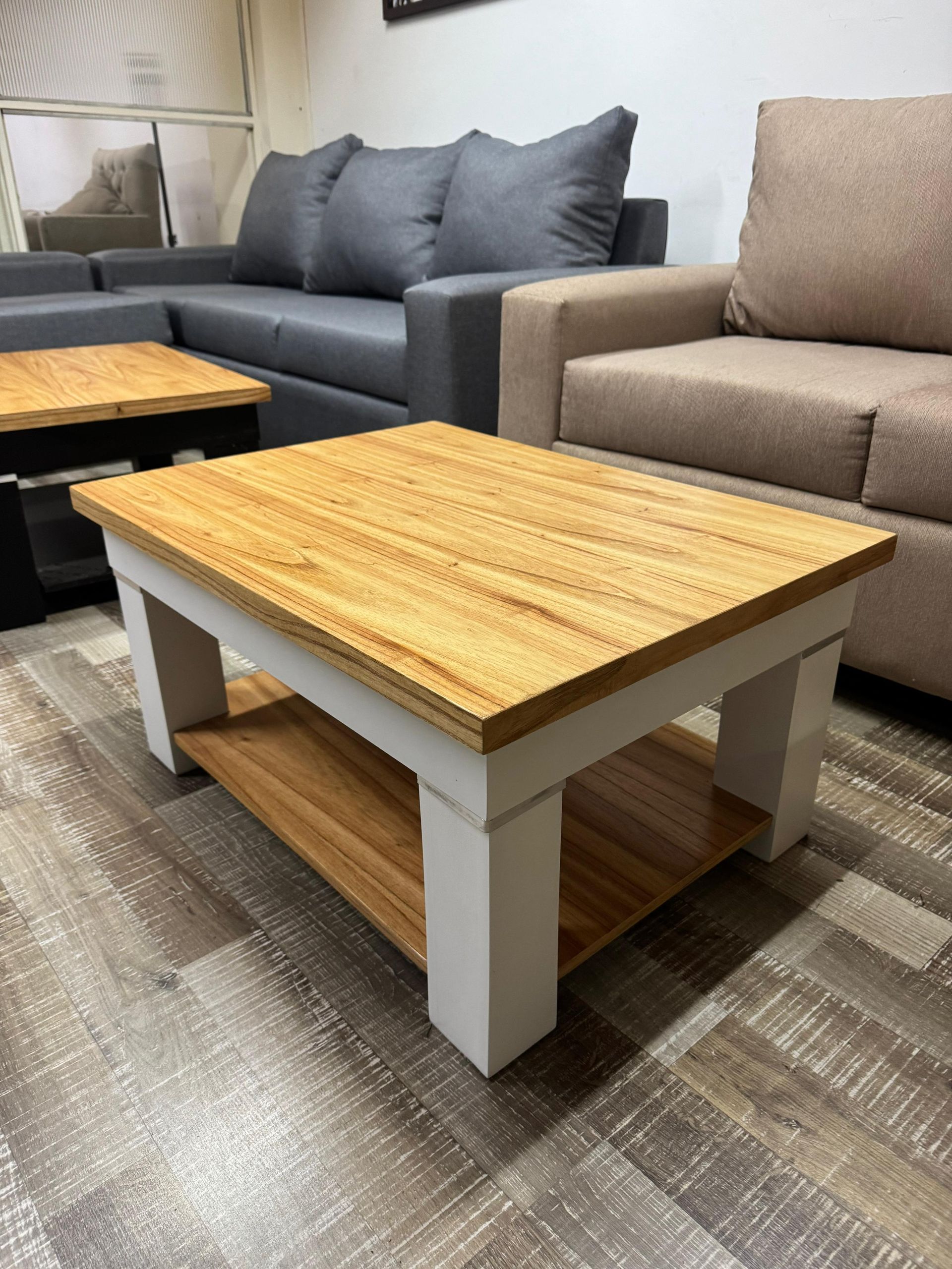 Una mesa de centro de madera con base blanca y estante inferior, colocada delante de sofás grises y beige