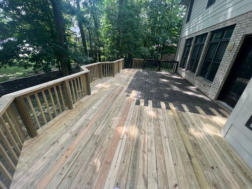 Wooden deck with railings beside a house, surrounded by trees and sunlight