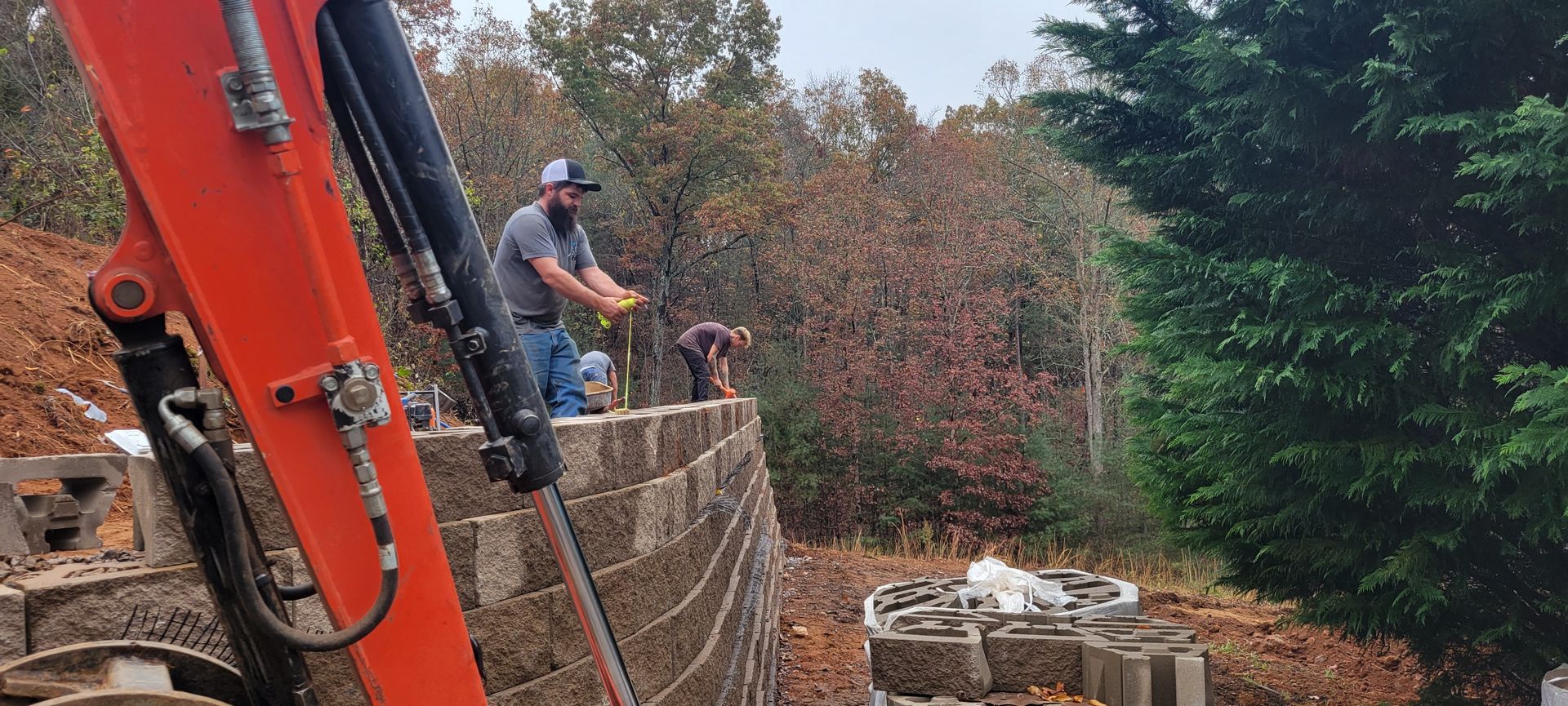 A man operating machinery, standing near a retaining wall. The scene is outdoors with trees.