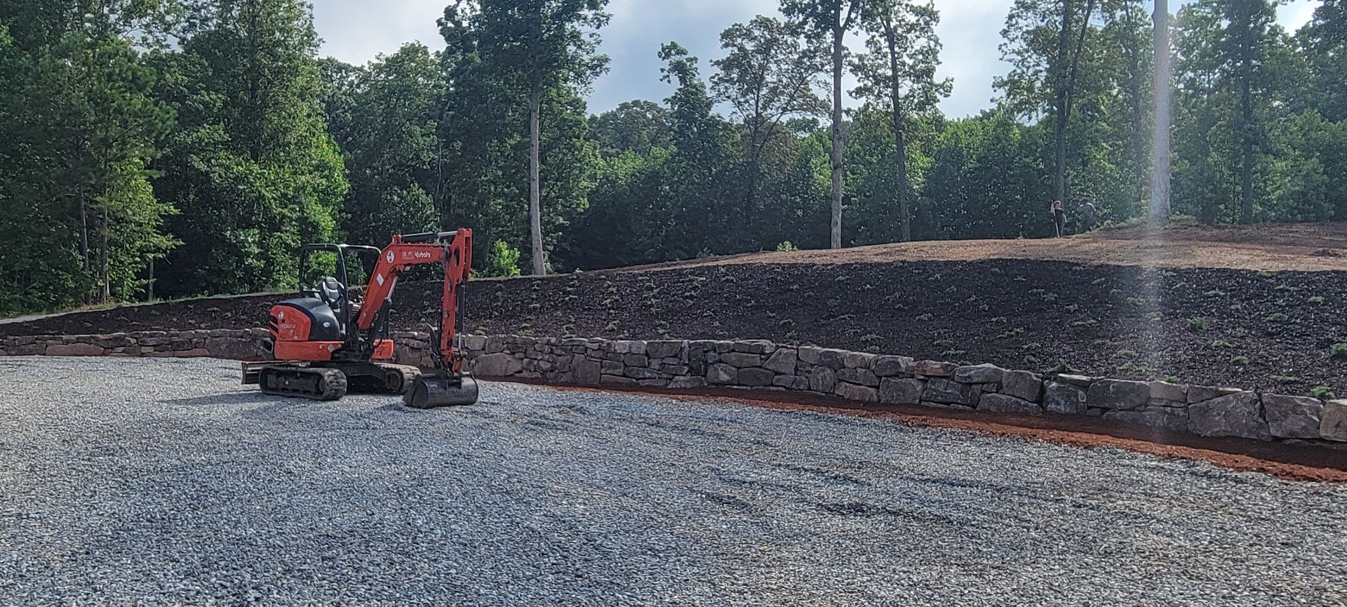 An orange excavator on a gravel bed near a stone wall in front of a forest.