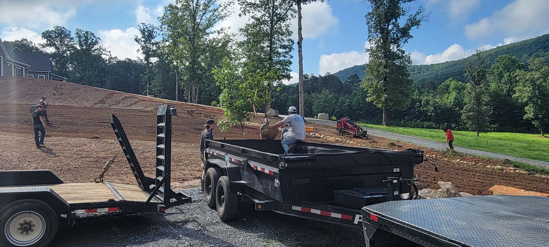Workers loading debris into a dump trailer on a hillside with trees and a mountain in the background.