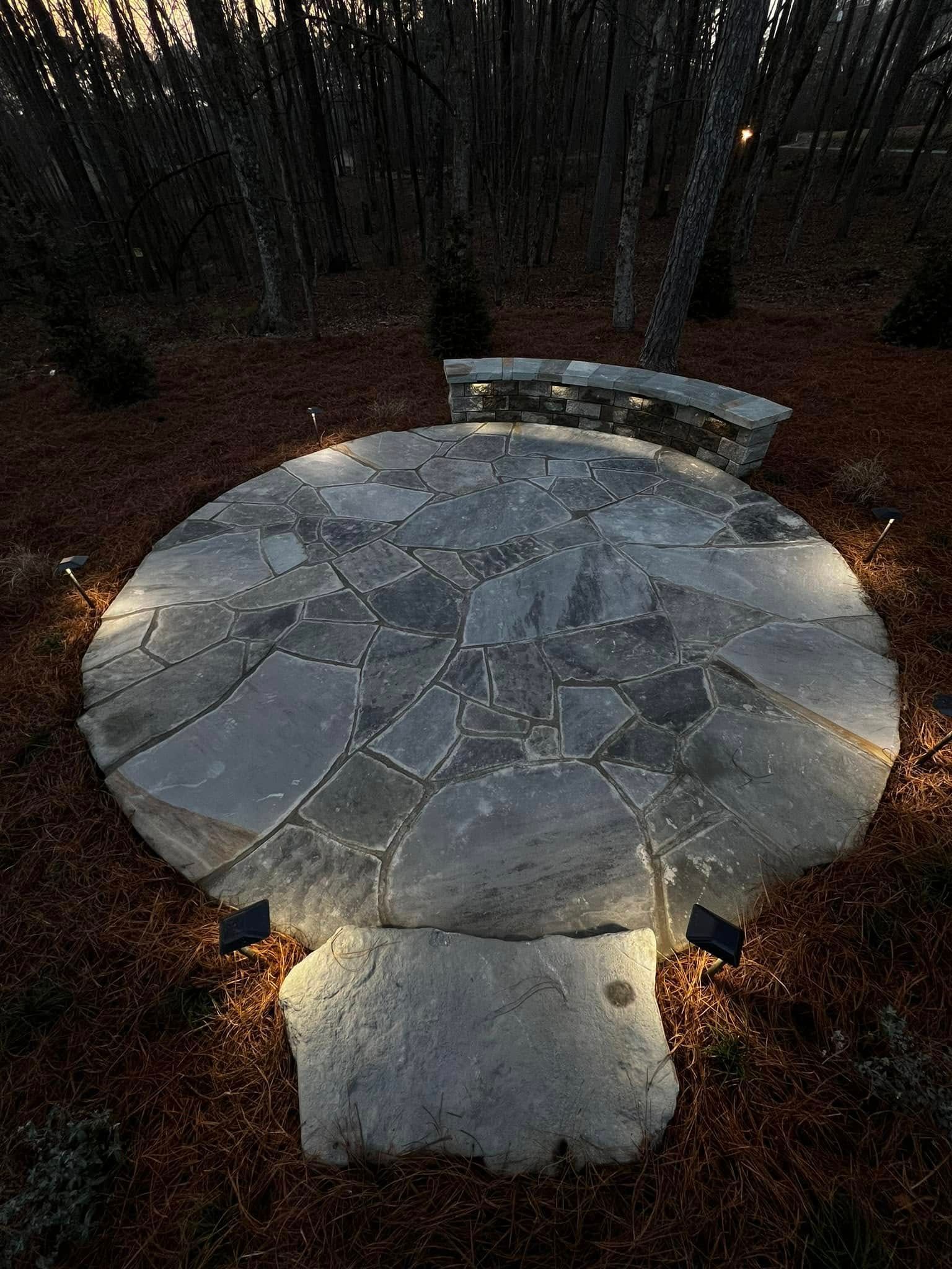 Stone patio with surrounding bench and ground lighting, set in a wooded area with pine straw.