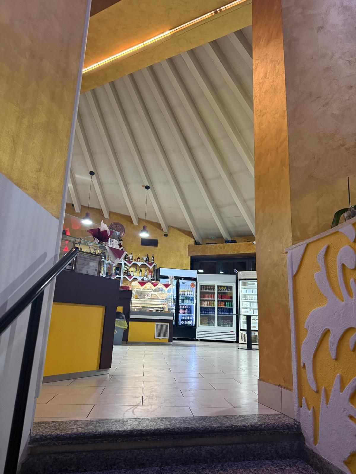 Ripresa interna dell'ingresso di un negozio con un bancone giallo brillante e refrigeratori per bevande visibili attraverso la porta.