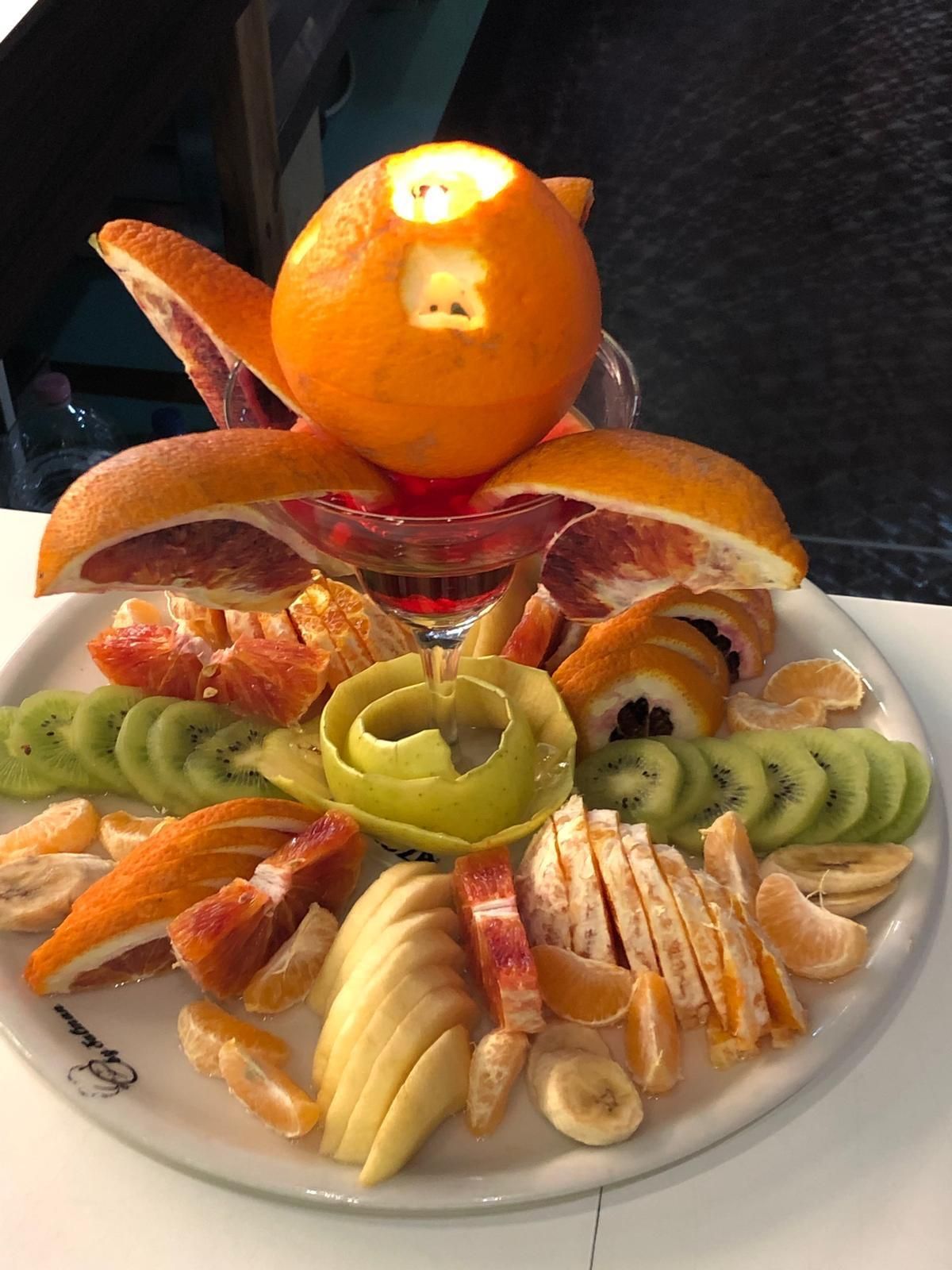 Vassoio di frutta con centrotavola di arancia intagliata, fette di agrumi, kiwi e spirale di mela su un piatto bianco.