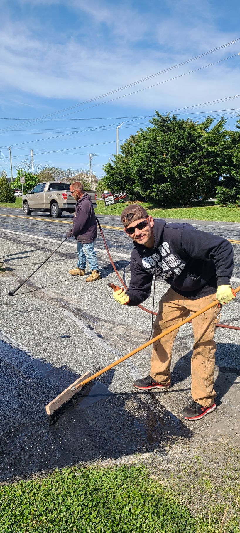 Two men cleaning a dark surface with long-handled brushes. Sunny day, one man smiles at camera.