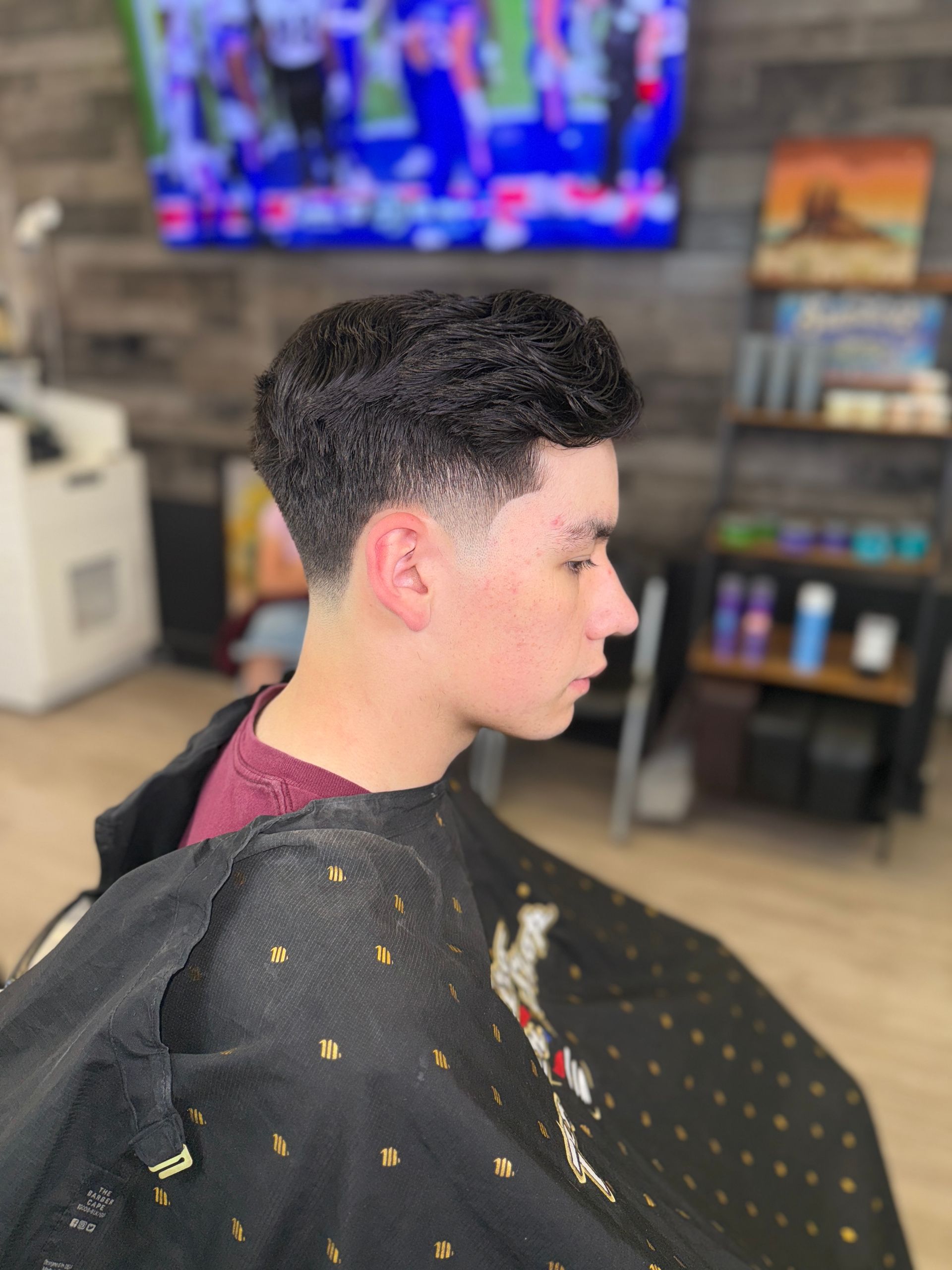 A person in a barber’s cape with a fresh skin fade haircut, sitting in a shop with a television screen in the background.